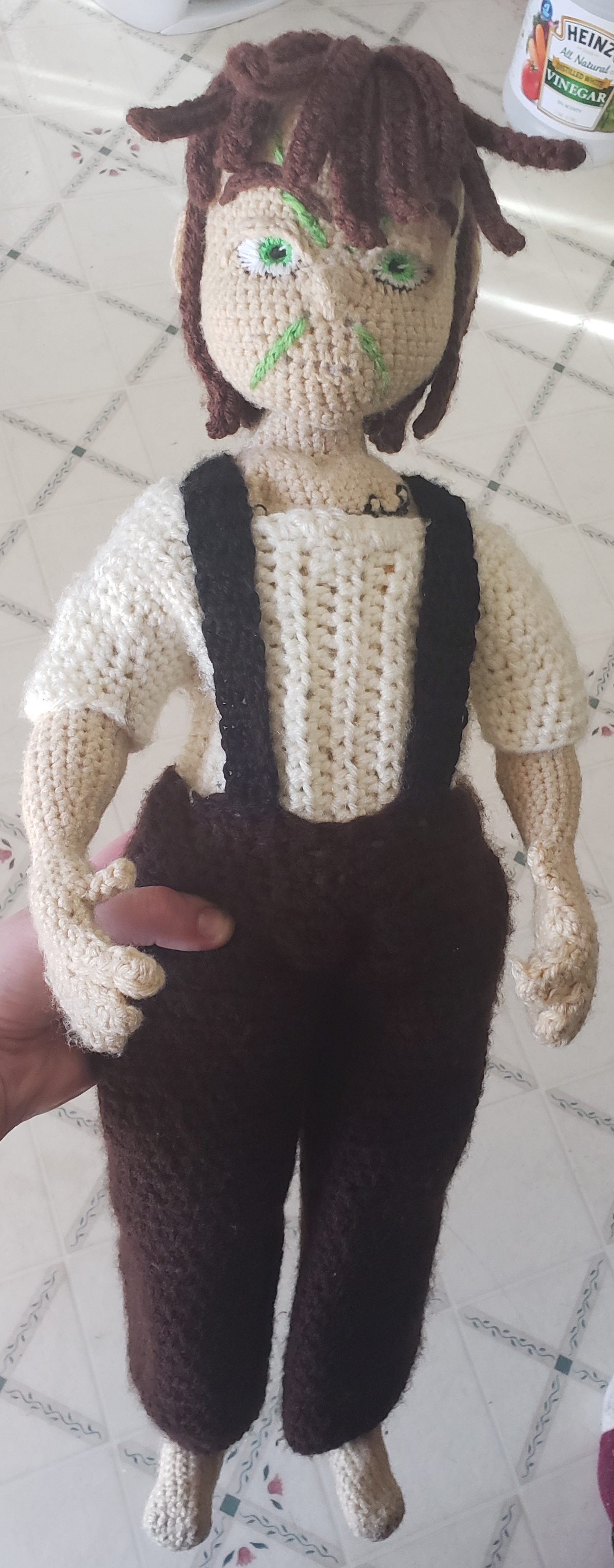 Crochet character doll with brown hair, embroidered green eyes and facial markings, a cream shirt, brown trousers, and suspenders.